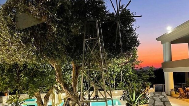 A pool area with stone planters, a wooden swing set, and a view of a building at dusk. - Blue Sky Villa in Crete (Hersonissos)