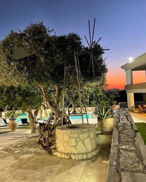 Un espace piscine agrémenté de jardinières en pierre, d’une balançoire en bois et offrant une vue sur un bâtiment au crépuscule.