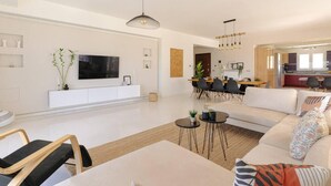 A modern living room with a sofa, coffee table, and TV. There is a dining area with chairs and a table. The room has a minimalist design with white walls and a large window. - Blue Sky Villa in Crete (Hersonissos)