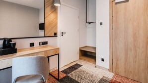 A modern hotel room with a wooden accent wall, a built-in desk with a coffee maker, a grey chair, a large mirror, and a wardrobe. - Hotel Vesper House (Gdansk)