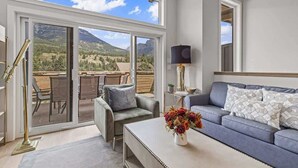 A modern living room with a large sliding glass door leading to a patio with outdoor furniture. There is a grey sofa with patterned cushions, a small table with a vase of flowers, and a floor lamp. - BRAND NEW|Vienna by SSR|TopView|Luxury|PRIVATE HOT TUB|4BR ( BL# RES-11679 ) (Canmore)