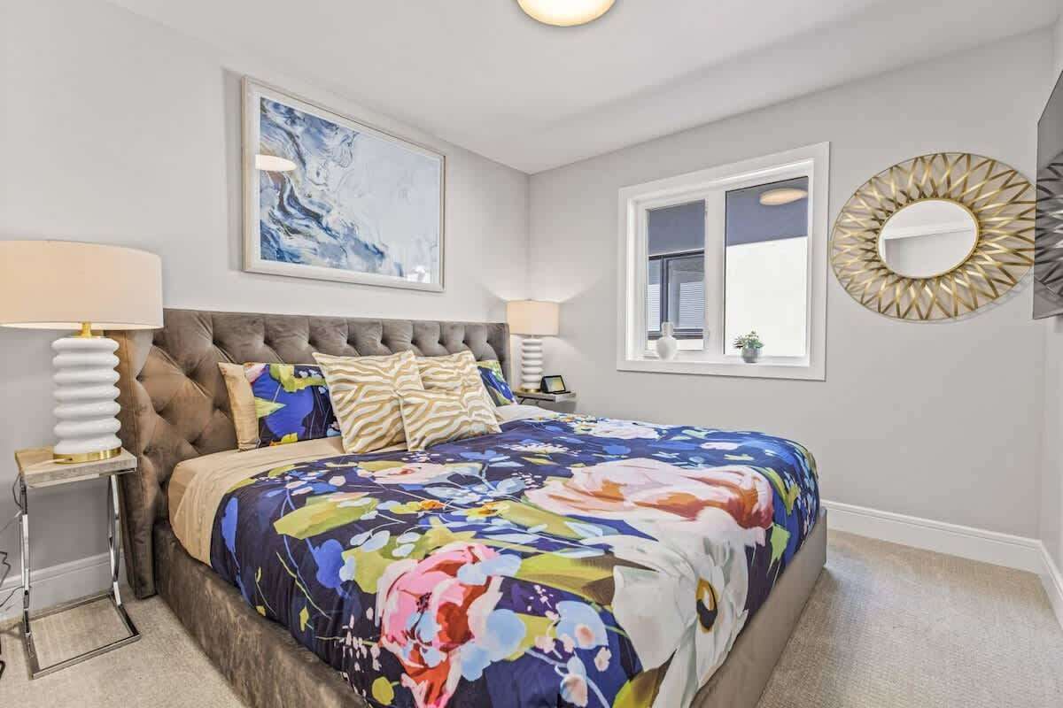 A bedroom with a tufted headboard, a floral bedspread, a large window with blinds, a round mirror, and a framed abstract painting.