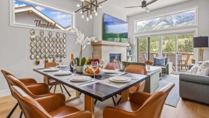 A dining area with a large table set for six, surrounded by leather chairs. A fireplace is visible in the background, and a large window offers a view of mountains. - BRAND NEW|Vienna by SSR|TopView|Luxury|PRIVATE HOT TUB|4BR ( BL# RES-11679 ) (Canmore)