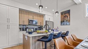 A modern kitchen with white and wooden cabinets, a central island with bar stools, and a dining area with a table set for four. - BRAND NEW|Vienna by SSR|TopView|Luxury|PRIVATE HOT TUB|4BR ( BL# RES-11679 ) (Canmore)