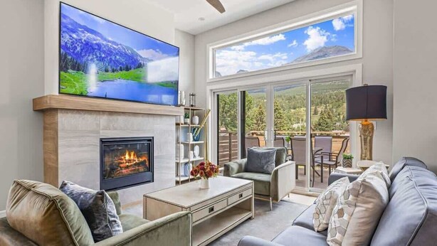 A living room with a fireplace, a large flat-screen TV, and a view of a mountain landscape through large windows. - BRAND NEW|Vienna by SSR|TopView|Luxury|PRIVATE HOT TUB|4BR ( BL# RES-11679 ) (Canmore)