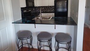 Cocina con gabinetes blancos, un refrigerador negro, una estufa negra y una encimera negra. Hay tres taburetes grises en la isla de la cocina.