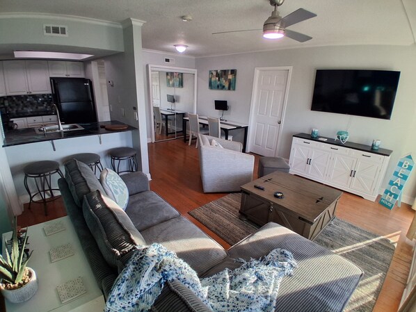 A modern living room with a sofa, a coffee table, and a TV. There is a kitchen area with a refrigerator and a dining table.
