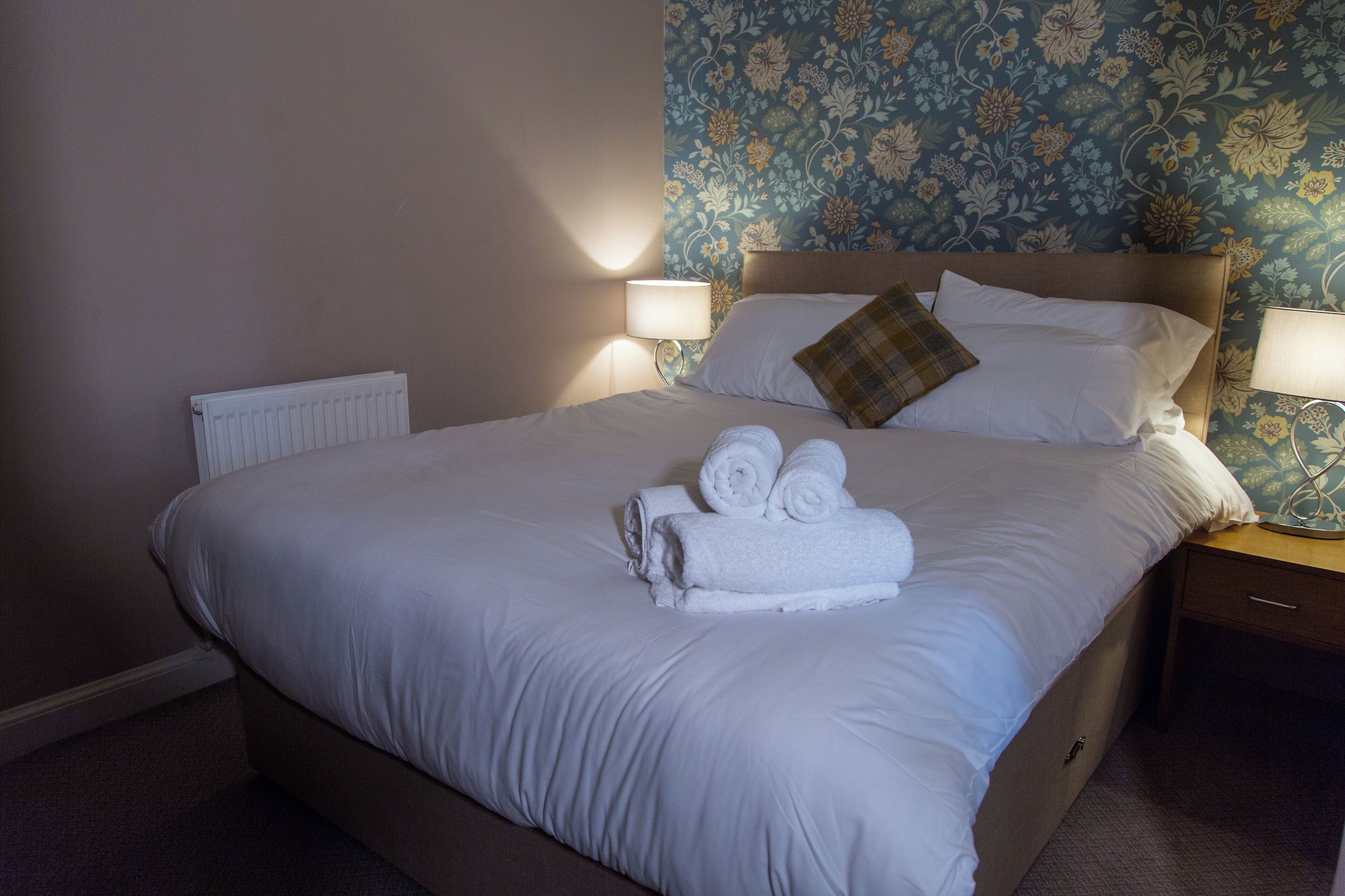 A neatly made bed with white linens and rolled towels, a wooden headboard, and a floral wallpaper pattern on the wall.