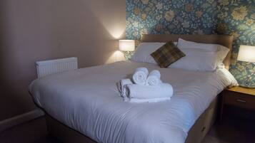 A neatly made bed with white linens and rolled towels, a wooden headboard, and a floral wallpaper pattern on the wall.