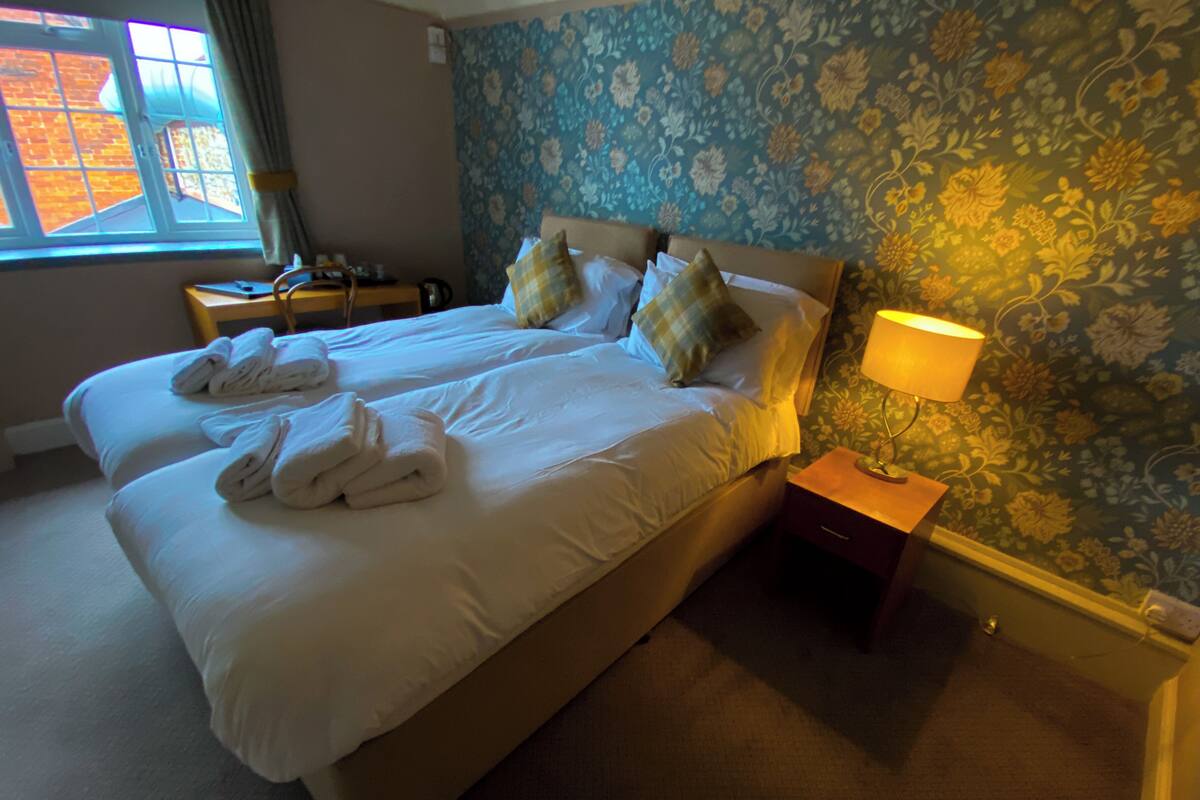 A hotel room with two beds, a nightstand with a lamp, and a window with curtains.