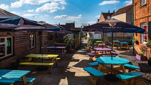 An outdoor patio area with multiple picnic tables and umbrellas, surrounded by brick buildings. - Market House (Glastonbury)