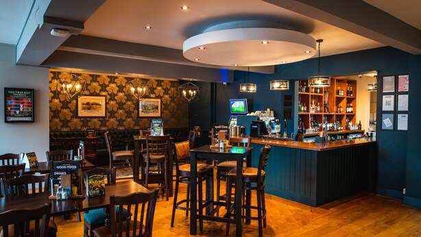 A restaurant with wooden tables and chairs, a bar area with shelves of bottles, and a wall with framed pictures and a menu board. - Market House (Glastonbury)