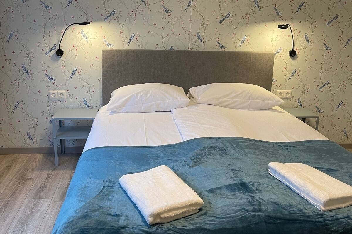A neatly made bed with a dark green bedspread, white pillows, and two folded towels on the bed. There are two bedside tables with lamps, and the wall features a bird and floral wallpaper pattern.