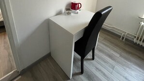 Ein kleiner, weißer Schreibtisch in L-Form mit einem schwarzen Stuhl, einer roten Tasse und einem weißen Gegenstand darauf.
