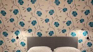 Ein ordentlich bezogenes Bett mit himbeerfarbener Bettdecke und weißen Kissen, ein grauer Kopfteil und eine Wand mit floralem Muster.