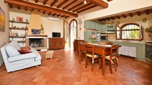 A spacious living area with a fireplace, wooden beams, a dining table, and a kitchen with green cabinets and a window. - Da Maria (Murlo)