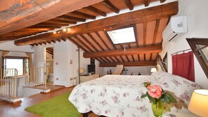 A bedroom with a wooden ceiling, a skylight, a bed with a floral bedspread, a green rug, a bedside table with a lamp, and a vase with a flower. - Da Maria (Murlo)
