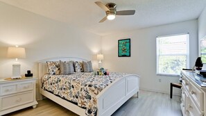 Une chambre à coucher comprenant un lit, des tables de chevet, une commode, un ventilateur de plafond, une fenêtre et une œuvre d’art murale.