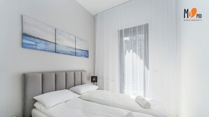 A modern bedroom with a large bed, a nightstand, a wall-mounted lamp, and a view of the outdoors through sheer curtains. - SUMMER LOFTS Apartamenty Premium (Ustronie Morskie)