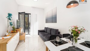 A modern living room with a sofa, a dining table set for two, and a large window. - SUMMER LOFTS Apartamenty Premium (Ustronie Morskie)