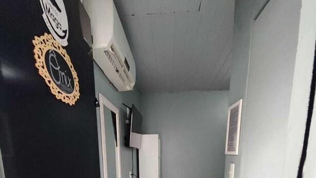Un pasillo estrecho con piso de baldosas blancas, una puerta blanca a la izquierda y una cama con manta gris al fondo.