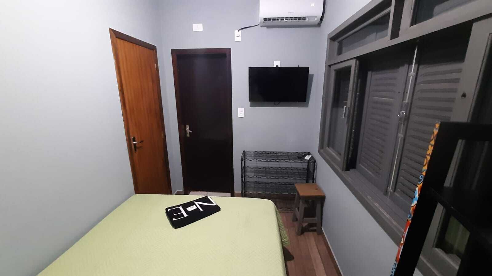 A small, narrow room with a bed, a TV mounted on the wall, a window with shutters, and an air conditioning unit.