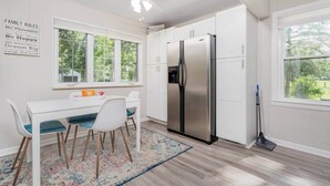 Une cuisine moderne avec des meubles blancs, un réfrigérateur en acier inoxydable, une table à manger avec des chaises et une grande fenêtre.