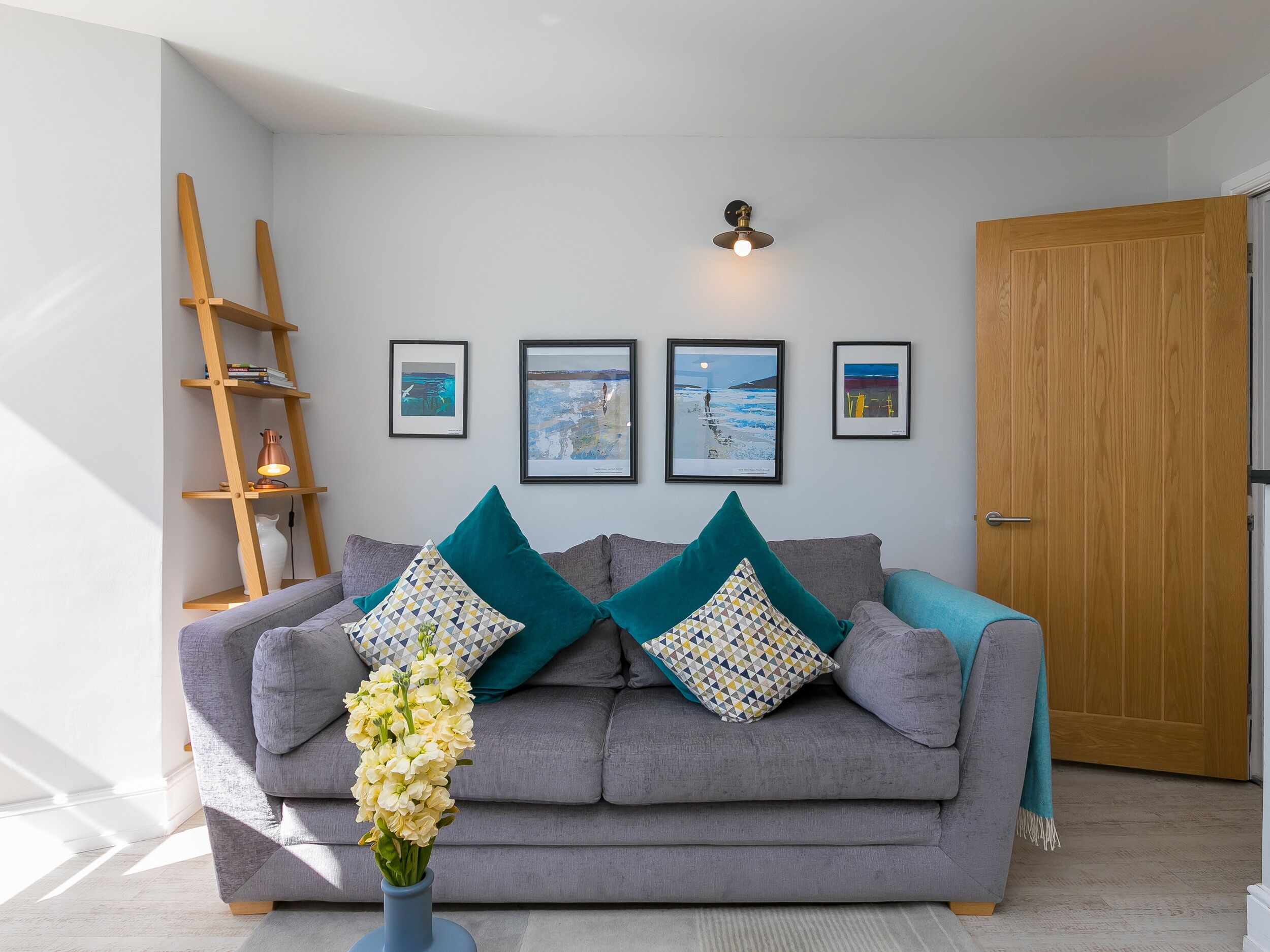 Ein Wohnzimmer mit grauem Sofa, türkisfarbenen Kissen, einer blauen Vase mit Blumen, einer Holzleiter und gerahmten Bildern an der Wand.