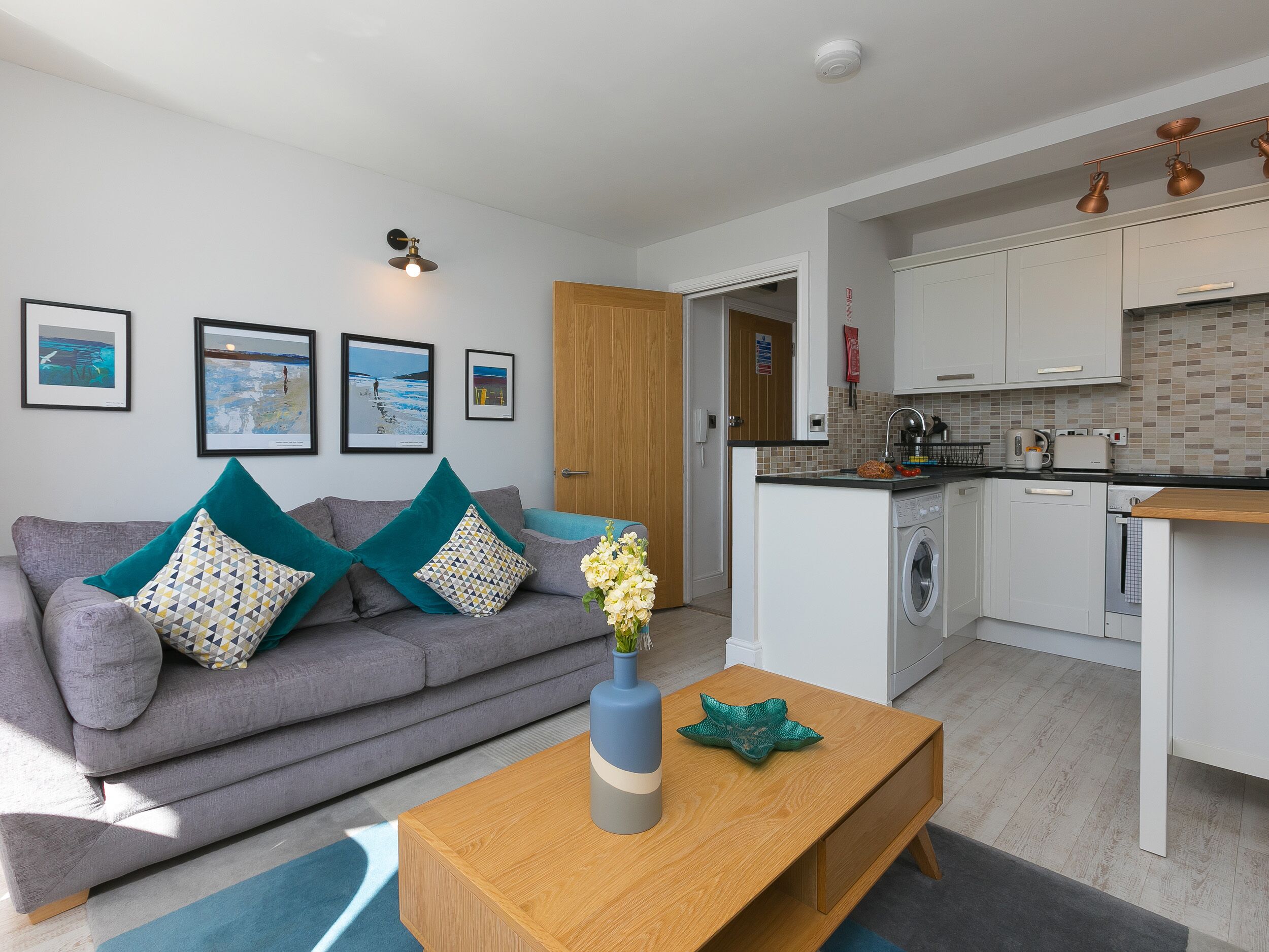 A modern living room with a grey sofa, teal cushions, and a wooden coffee table with a vase of flowers. Adjacent is a compact kitchen with white cabinets and a tiled backsplash.