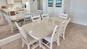 A modern dining area with a glass table and white chairs, a kitchen in the background, and a door with a beach mural. - Oceanfront 3 Bedroom Condo - Steps to the beach and ocean. Perfecty located on 119th Street! Sandy Square 304 (Ocean City)
