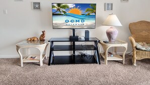A living room with a TV displaying a beach scene, a small table with a lamp, and a wicker chair. - Oceanfront 3 Bedroom Condo - Steps to the beach and ocean. Perfecty located on 119th Street! Sandy Square 304 (Ocean City)