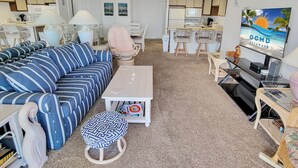 A modern living room with a sofa, coffee table, and ottoman. There is a kitchen area with a microwave, stove, and dining table. A television is mounted on the wall. - Oceanfront 3 Bedroom Condo - Steps to the beach and ocean. Perfecty located on 119th Street! Sandy Square 304 (Ocean City)