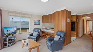 A compact living space with a TV, two leather armchairs, a wooden coffee table, a kitchenette, and a view of a cityscape. - Views on Ballingalla (Narooma)