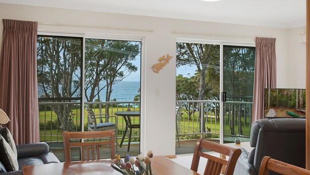 A room with a view of the sea, a dining area with a table and chairs, and a large window with curtains. - Views on Ballingalla (Narooma)