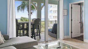 Una habitación con puerta corrediza de vidrio que da a un balcón con vista al océano y palmeras. Hay una silla y una mesa con un control remoto.