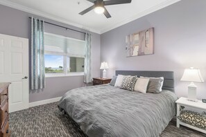 Une chambre avec un grand lit, un ventilateur de plafond, une fenêtre avec des rideaux, une table de chevet avec une lampe et une porte.