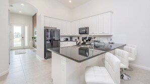 A modern kitchen with white cabinets, a large island with bar stools, stainless steel appliances, and a view to the outside. - Last Minute Disney Getaway! Private Pool, Hot Tub & Game Room. Book Now! #41843 (Orlando)