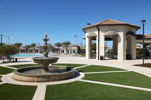 A fountain with multiple tiers, a gazebo, and a pool in a landscaped area with green grass and pathways.