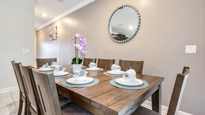 Zona de dinar amb taula de fusta preparada per a sis, envoltada de cadires de fusta. Hi ha un vas amb flors morades sobre la taula, i un mirall rodó amb decoracions similars a perles penjat a la paret.
