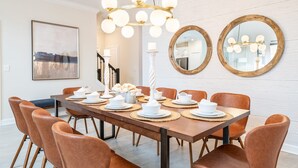 Une salle à manger avec une longue table en bois, dressée pour six personnes, entourée de six chaises en cuir marron. Deux miroirs ronds sont accrochés au mur, ainsi qu’un lustre à plusieurs lampes et un tableau encadré au-dessus de la table.