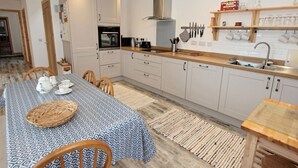Une cuisine moderne avec des armoires blanches, un four encastré et une hotte de cuisinière en acier inoxydable. On y trouve également un coin repas avec une nappe à motifs et des chaises.