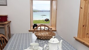Une table recouverte d’une nappe à motifs, une théière blanche, des tasses et un panier en osier. Une fenêtre offre une vue sur une étendue d’eau et une zone herbeuse où se trouvent des personnes et un véhicule.