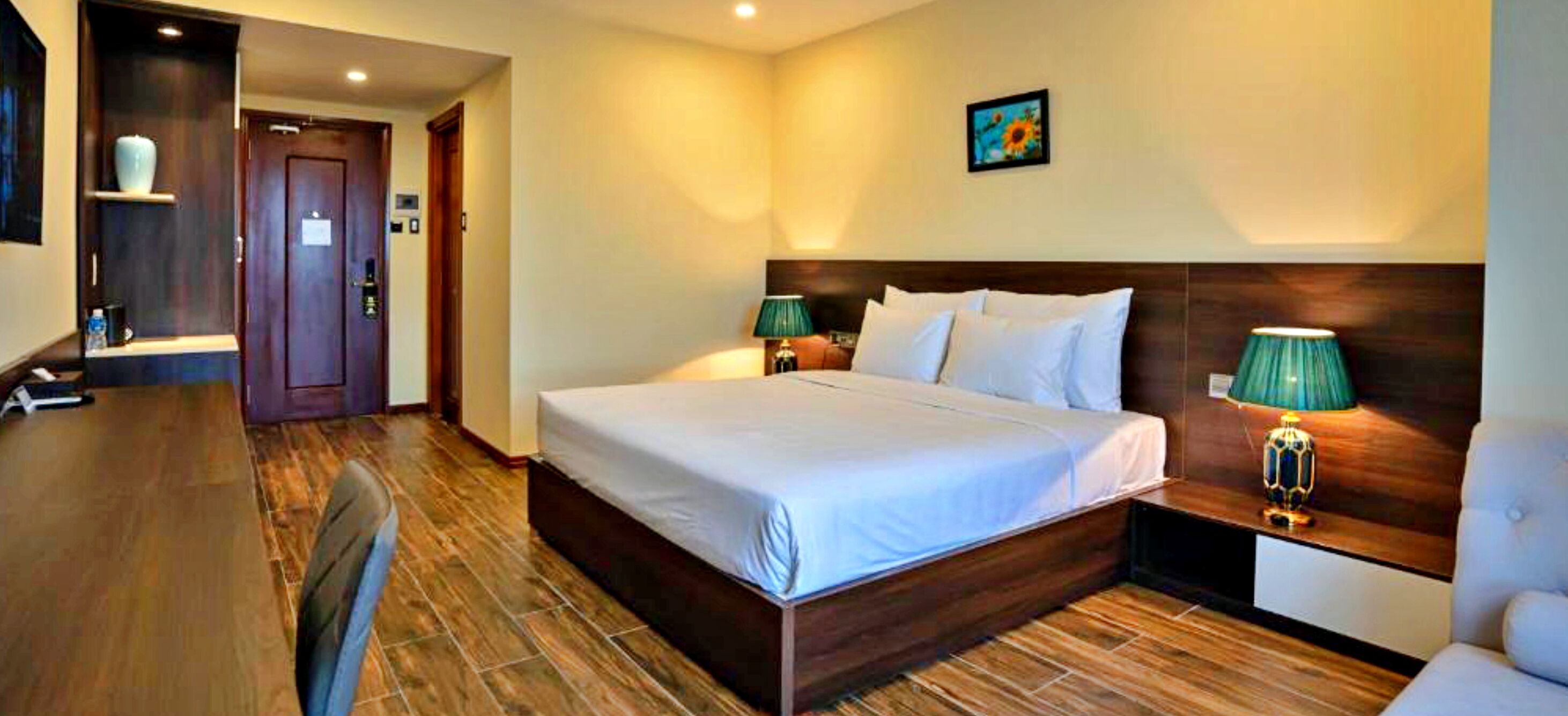 Quarto de hotel com cama, abajures, cabeceira de madeira, escrivaninha e uma cadeira.