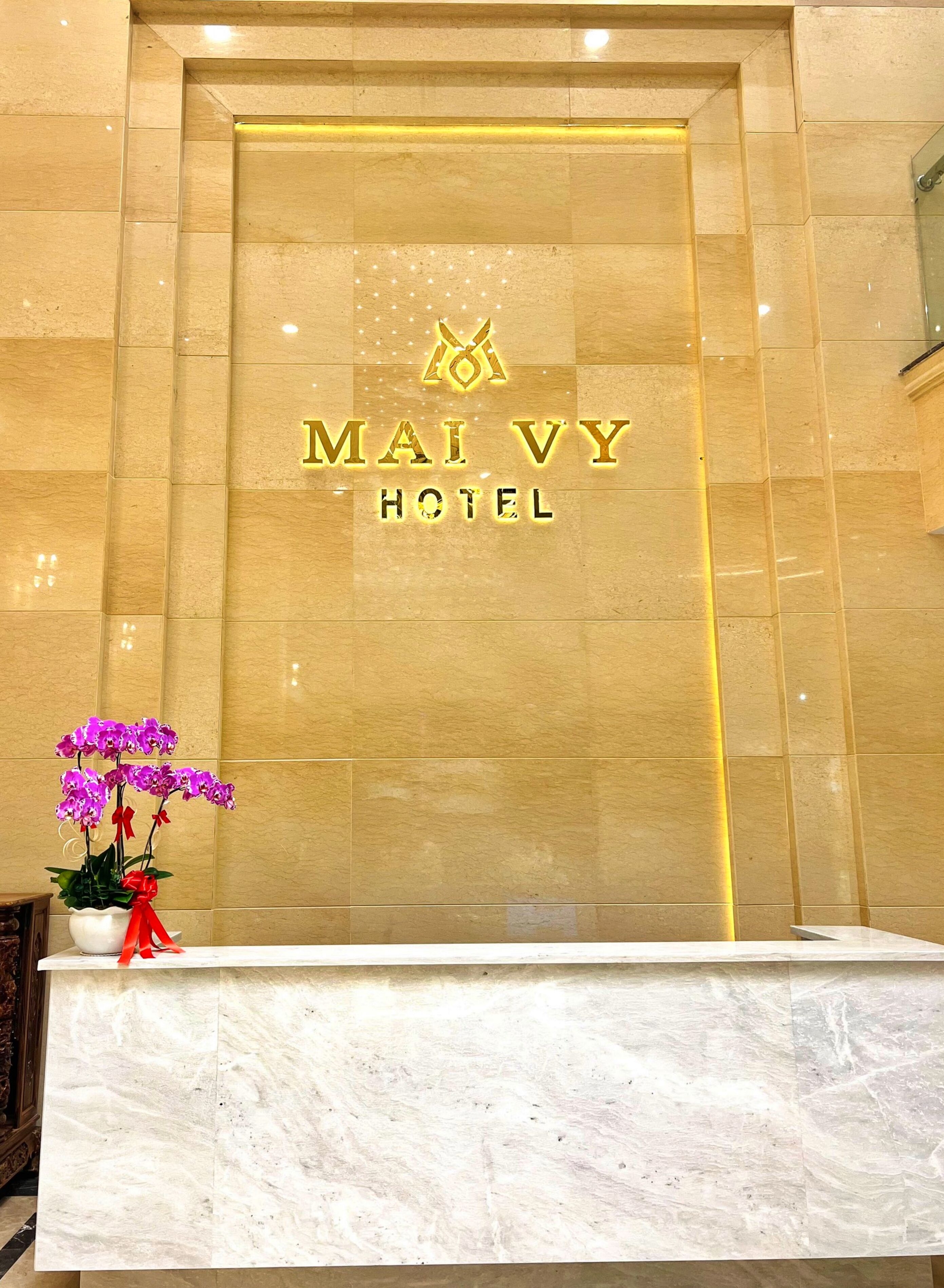 A reception desk with a marble top, a potted orchid, and the hotel name 'MAI VY HOTEL' illuminated on the wall.