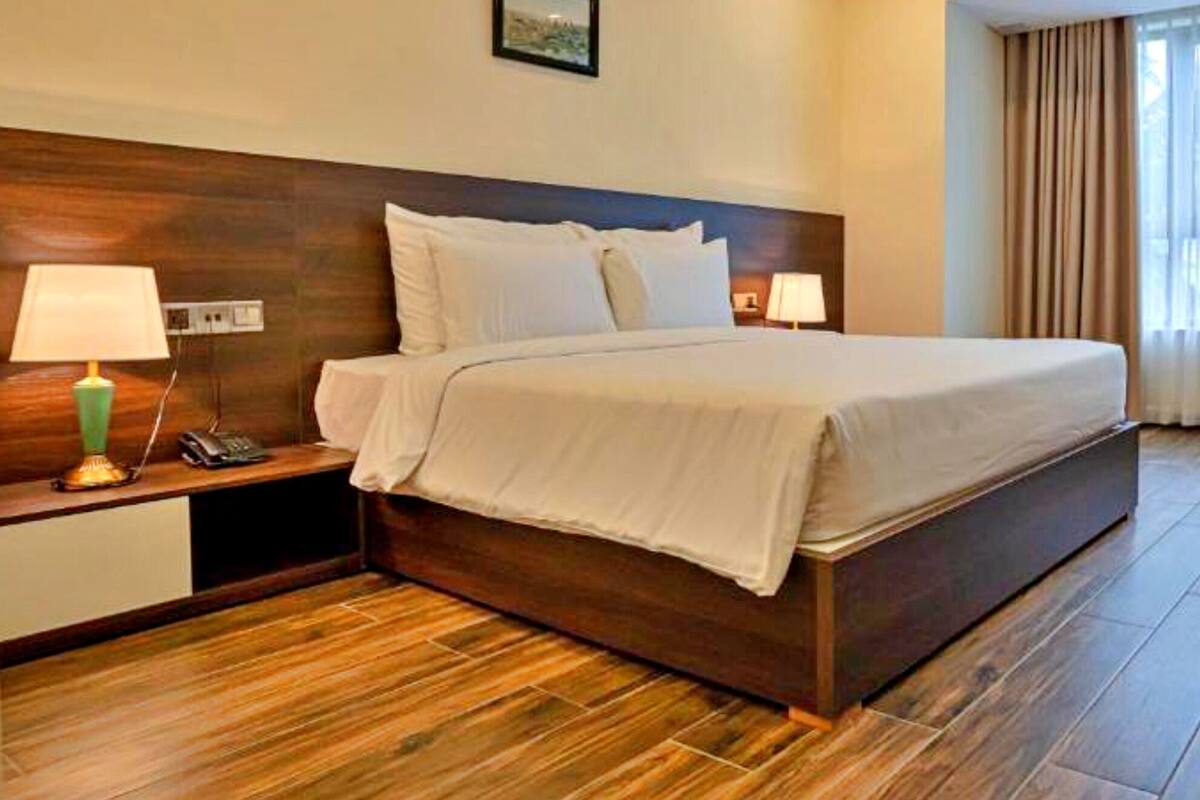 Une chambre d’hôtel avec un grand lit, un plancher en bois, un bureau avec une lampe, une télévision et une fenêtre avec des rideaux.