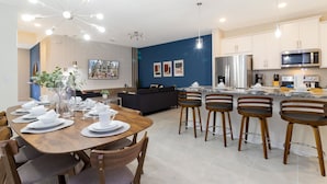 Cozinha moderna com ilha central, eletrodomésticos de aço inoxidável e área de jantar com mesa de madeira e banquetas.