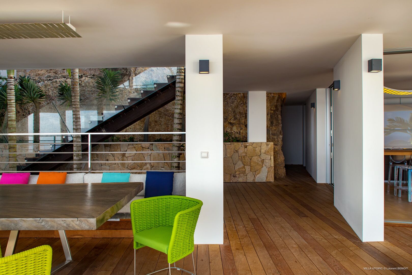 A modern interior with a wooden floor, a staircase, and a dining area with colorful cushions.