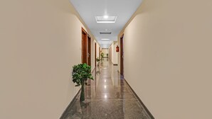 Um corredor longo e bem iluminado, com portas de madeira em ambos os lados, piso polido e plantas em vasos ao longo das paredes.
