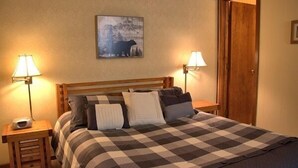Een slaapkamer met een geruit beddegoed, twee bedlampen, een houten hoofdeinde en een schilderij van een beer aan de muur.