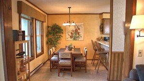 Een eetruimte met een houten tafel en stoelen, een kroonluchter en een groot raam met zonwering.